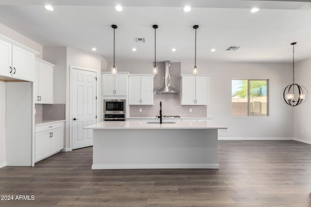 kitchen with white cabinets, hanging light fixtures, wall chimney exhaust hood, built in microwave, and an island with sink