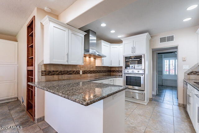 kitchen featuring appliances with stainless steel finishes, tasteful backsplash, white cabinets, kitchen peninsula, and wall chimney exhaust hood