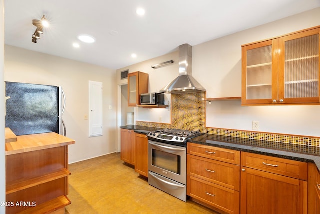 kitchen with track lighting, wall chimney exhaust hood, stainless steel appliances, dark stone counters, and decorative backsplash