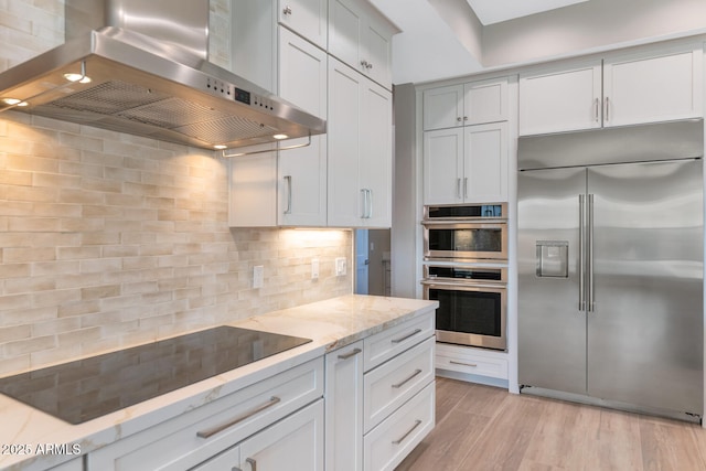 kitchen featuring tasteful backsplash, light stone countertops, light wood-type flooring, appliances with stainless steel finishes, and wall chimney exhaust hood