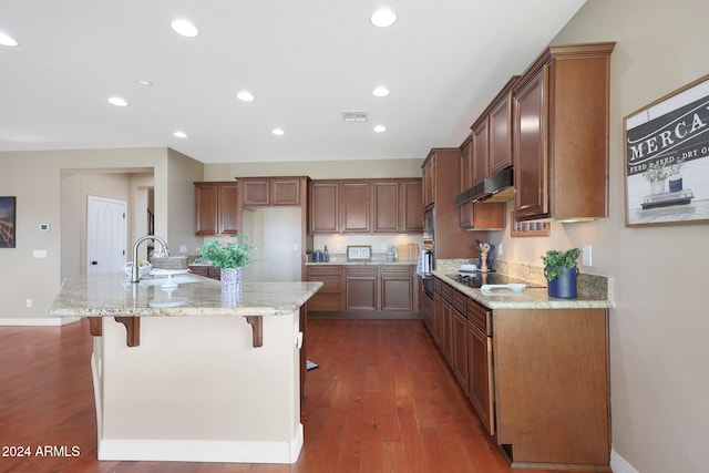 kitchen with a kitchen island with sink, sink, light stone countertops, a breakfast bar, and dark hardwood / wood-style flooring