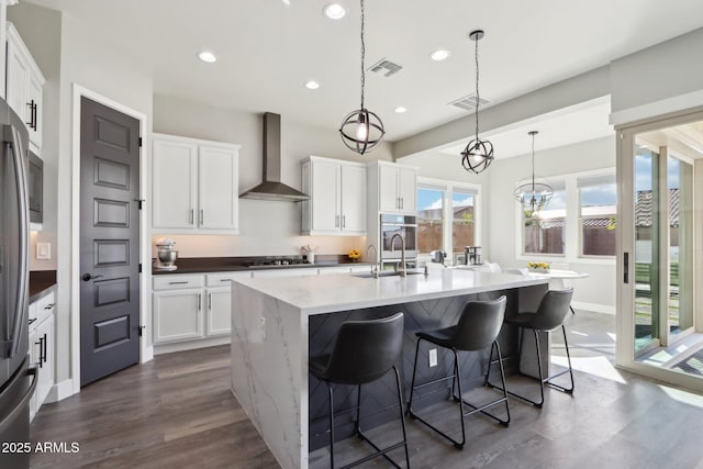 kitchen with hanging light fixtures, white cabinetry, a kitchen island with sink, and wall chimney range hood