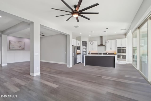 kitchen with dark wood-type flooring, wall chimney exhaust hood, decorative light fixtures, a kitchen island with sink, and white cabinets