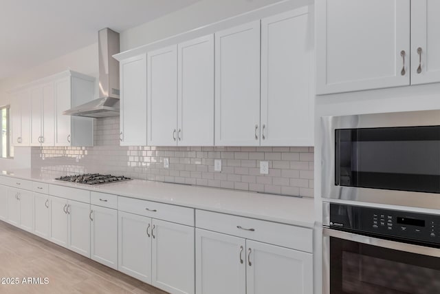 kitchen featuring appliances with stainless steel finishes, white cabinetry, wall chimney range hood, tasteful backsplash, and light hardwood / wood-style floors