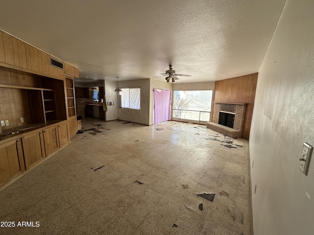 unfurnished living room featuring visible vents, a brick fireplace, ceiling fan, wooden walls, and a textured ceiling