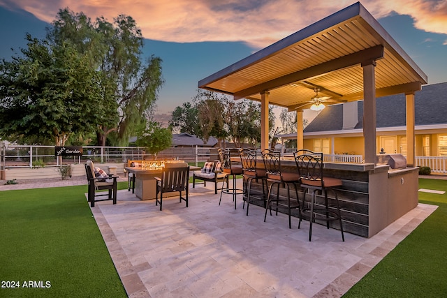 patio terrace at dusk featuring a lawn, ceiling fan, an outdoor fire pit, grilling area, and a bar