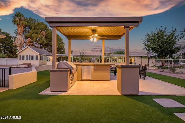 patio terrace at dusk featuring an outdoor bar, ceiling fan, a yard, area for grilling, and exterior kitchen