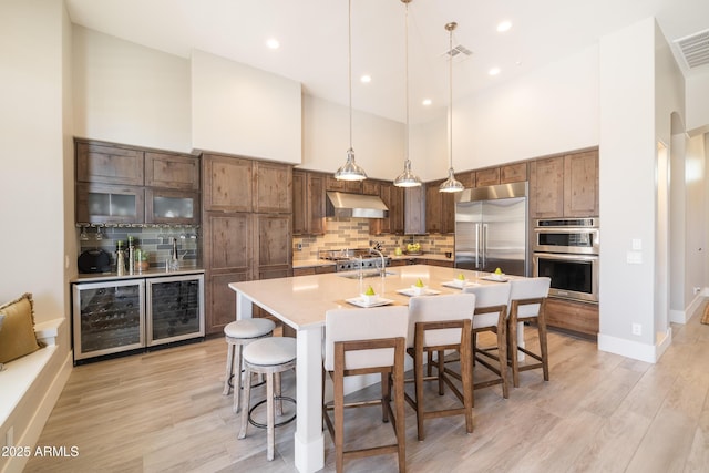 kitchen with under cabinet range hood, a towering ceiling, appliances with stainless steel finishes, a kitchen breakfast bar, and tasteful backsplash