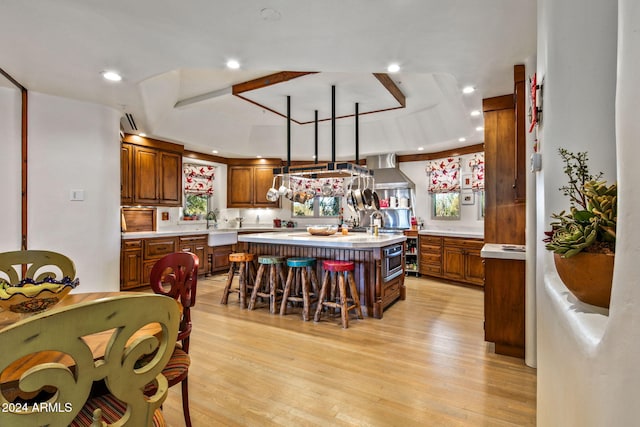 kitchen with wall chimney range hood, an island with sink, light hardwood / wood-style flooring, and a healthy amount of sunlight