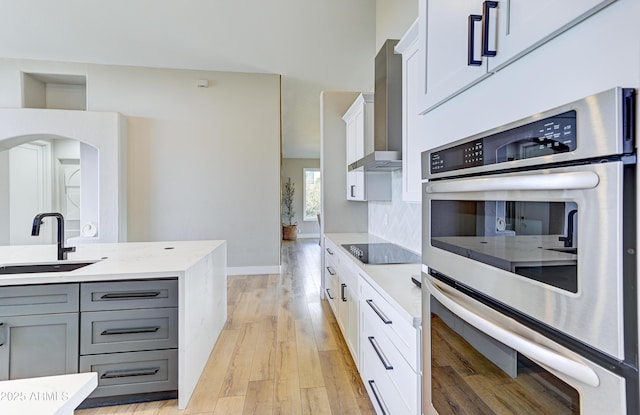 kitchen featuring double oven, gray cabinetry, a sink, white cabinetry, and wall chimney exhaust hood