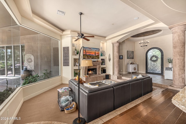 living room featuring ornate columns, ceiling fan with notable chandelier, a raised ceiling, dark hardwood / wood-style floors, and a tiled fireplace