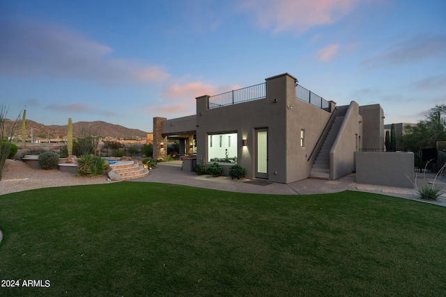 back house at dusk with a mountain view, a balcony, a lawn, and a patio