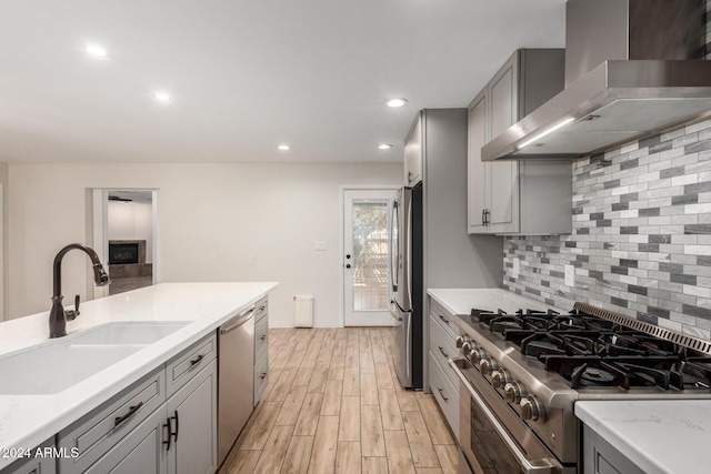 kitchen with appliances with stainless steel finishes, wall chimney exhaust hood, sink, light hardwood / wood-style flooring, and gray cabinets