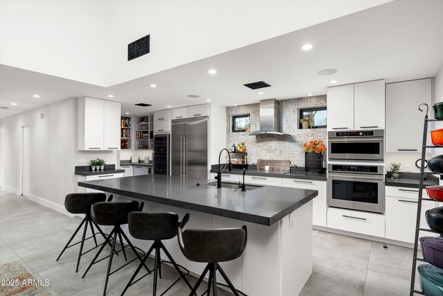 kitchen featuring sink, wall chimney exhaust hood, an island with sink, appliances with stainless steel finishes, and white cabinetry