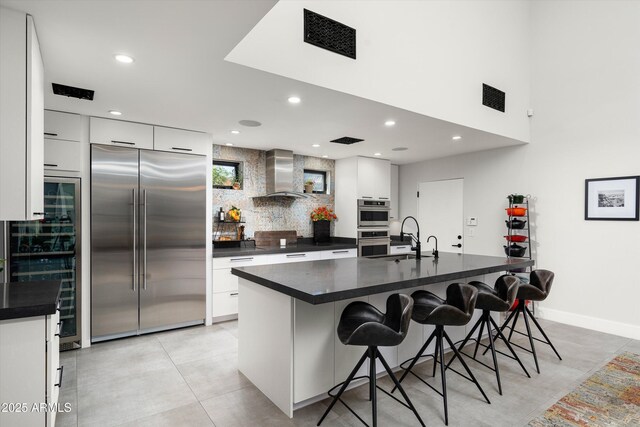 kitchen with a kitchen breakfast bar, wall chimney exhaust hood, stainless steel appliances, a kitchen island with sink, and white cabinets