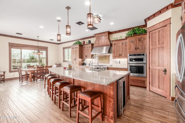 kitchen with light wood finished floors, custom range hood, appliances with stainless steel finishes, and brown cabinetry