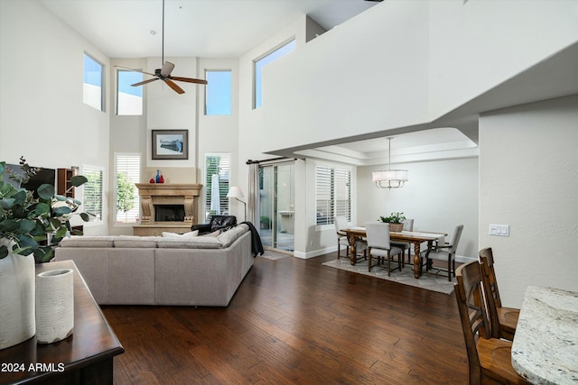 living area featuring a fireplace with raised hearth, dark wood-type flooring, ceiling fan with notable chandelier, and baseboards