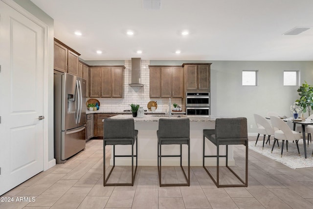 kitchen featuring a kitchen island with sink, a kitchen breakfast bar, wall chimney exhaust hood, light stone countertops, and stainless steel appliances