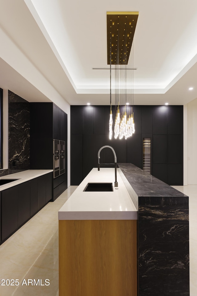 kitchen with pendant lighting, a tray ceiling, a kitchen island with sink, and sink