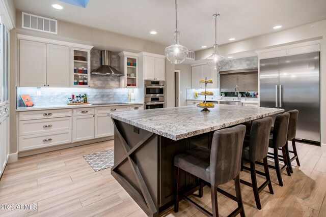 kitchen featuring light hardwood / wood-style floors, tasteful backsplash, wall chimney range hood, appliances with stainless steel finishes, and a large island