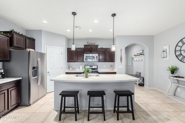 kitchen with light tile patterned floors, a kitchen island with sink, hanging light fixtures, stainless steel appliances, and dark brown cabinetry