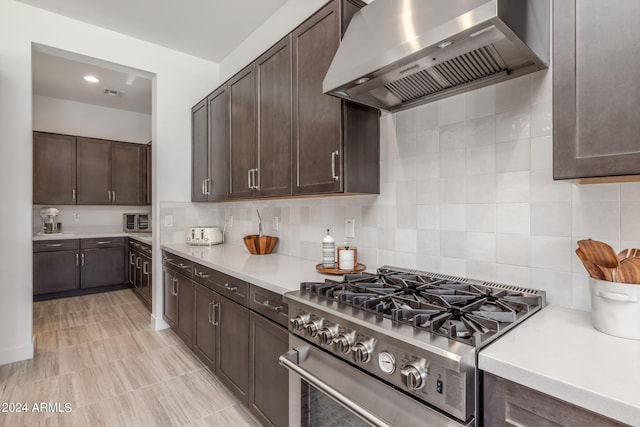 kitchen featuring stainless steel stove, wall chimney exhaust hood, backsplash, dark brown cabinetry, and light tile flooring