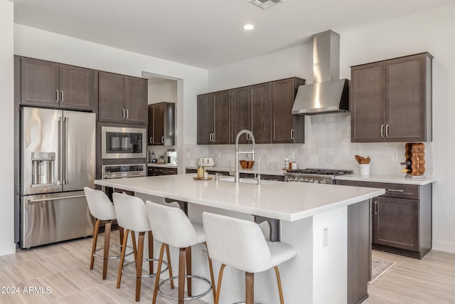 kitchen featuring a kitchen breakfast bar, a center island with sink, backsplash, wall chimney exhaust hood, and appliances with stainless steel finishes