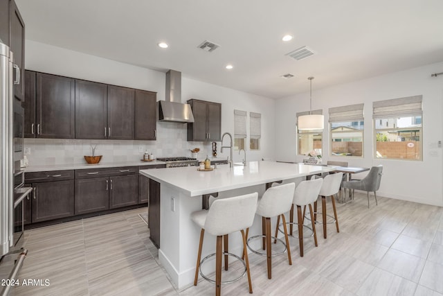 kitchen with hanging light fixtures, a kitchen island with sink, wall chimney exhaust hood, tasteful backsplash, and light tile floors