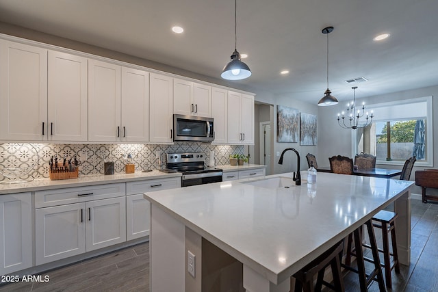 kitchen with white cabinets, appliances with stainless steel finishes, decorative light fixtures, dark wood-type flooring, and a kitchen island with sink