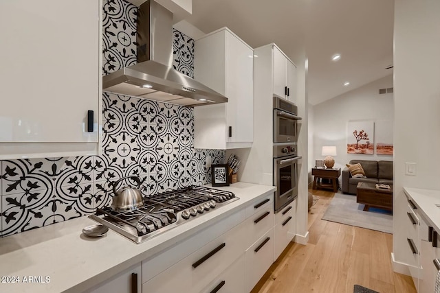 kitchen featuring white cabinetry, wall chimney exhaust hood, light hardwood / wood-style flooring, vaulted ceiling, and appliances with stainless steel finishes