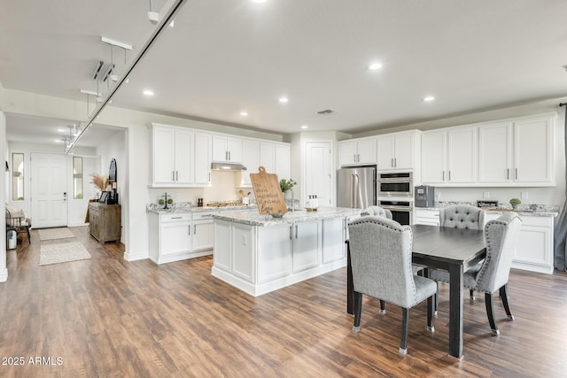 kitchen with light stone countertops, a kitchen island with sink, stainless steel appliances, under cabinet range hood, and white cabinetry