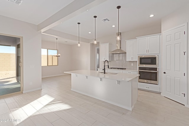 kitchen featuring sink, stainless steel appliances, wall chimney exhaust hood, white cabinets, and a center island with sink