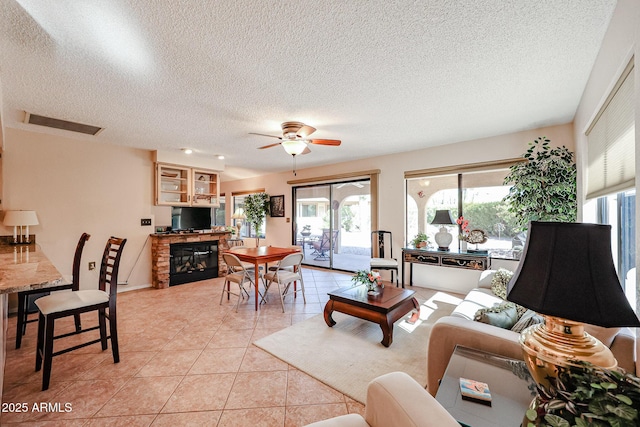 tiled living room featuring ceiling fan, a stone fireplace, and a textured ceiling