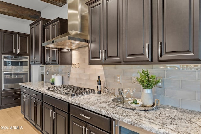 kitchen with tasteful backsplash, wall chimney range hood, appliances with stainless steel finishes, light wood-type flooring, and beamed ceiling