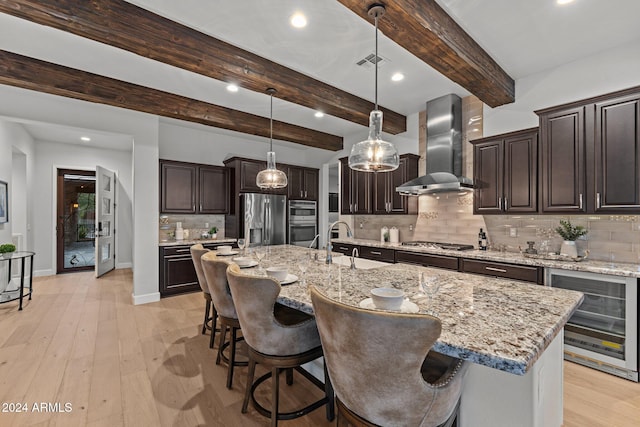 kitchen featuring wall chimney range hood, stainless steel appliances, beamed ceiling, wine cooler, and a kitchen island with sink