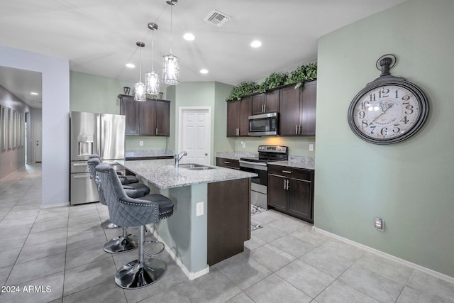 kitchen with light stone countertops, appliances with stainless steel finishes, dark brown cabinets, a breakfast bar area, and a center island with sink