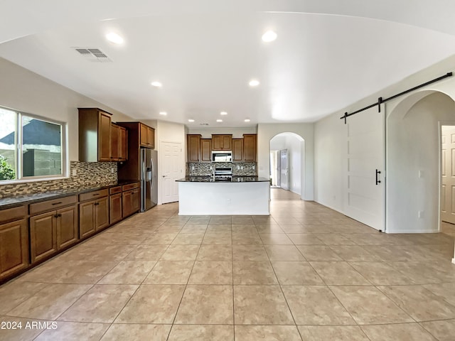 kitchen featuring a barn door, light tile patterned floors, an island with sink, tasteful backsplash, and stainless steel appliances