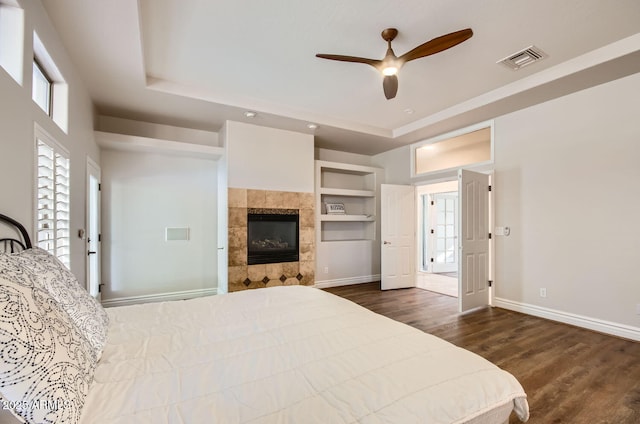 bedroom with a tiled fireplace, dark hardwood / wood-style floors, a raised ceiling, and ceiling fan