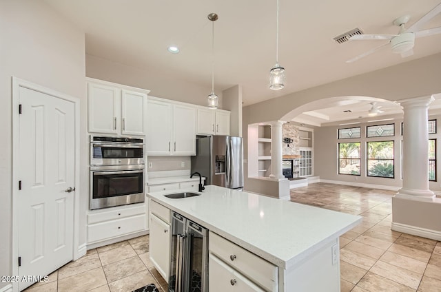 kitchen featuring wine cooler, an island with sink, ceiling fan, stainless steel appliances, and decorative columns