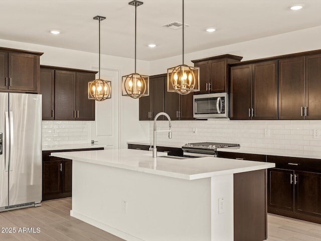 kitchen featuring stainless steel appliances, an island with sink, backsplash, dark brown cabinetry, and pendant lighting