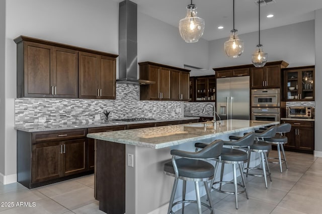 kitchen featuring light stone counters, appliances with stainless steel finishes, glass insert cabinets, a sink, and wall chimney range hood