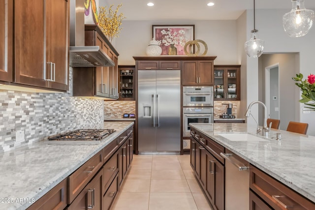 kitchen featuring light tile patterned floors, stainless steel appliances, a sink, wall chimney range hood, and decorative light fixtures