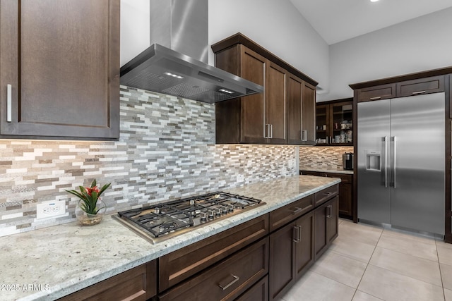 kitchen with light stone counters, stainless steel appliances, dark brown cabinets, wall chimney range hood, and tasteful backsplash