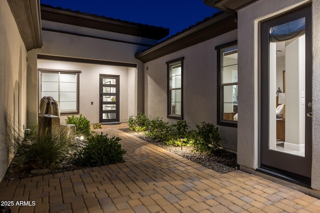 exterior entry at night with a tile roof, a patio area, and stucco siding