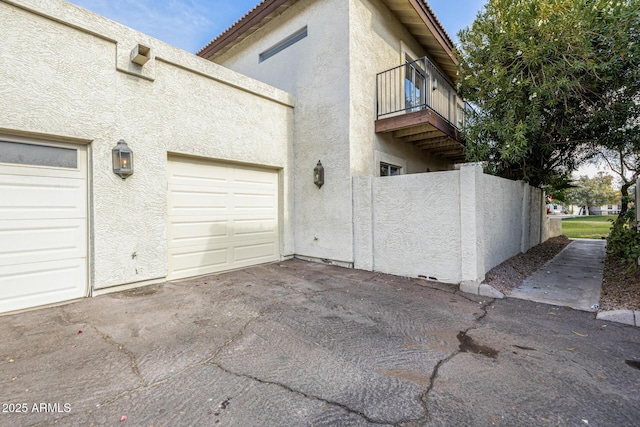 view of home's exterior featuring driveway, a balcony, an attached garage, fence, and stucco siding