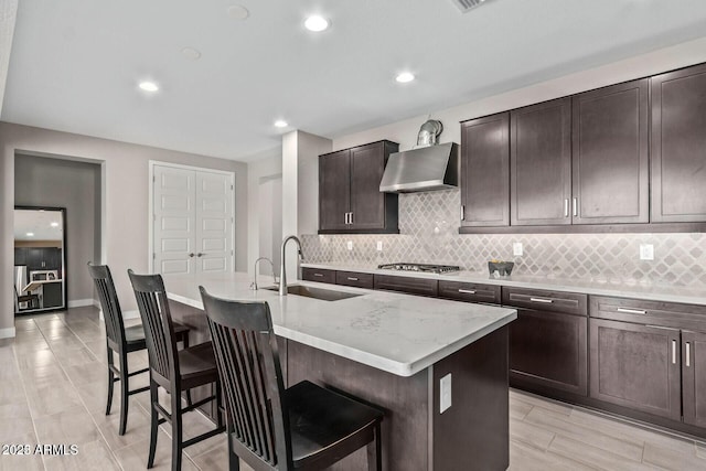 kitchen featuring a center island with sink, stainless steel gas cooktop, a breakfast bar, a sink, and wall chimney range hood