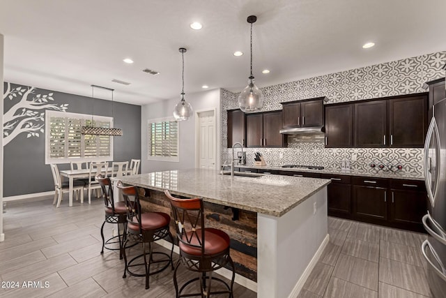 kitchen featuring a center island with sink, a kitchen breakfast bar, sink, hanging light fixtures, and light stone countertops