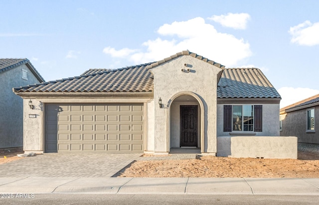 mediterranean / spanish house with a garage, a tiled roof, decorative driveway, and stucco siding