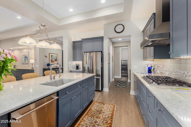 kitchen with stainless steel appliances, sink, light hardwood / wood-style flooring, hanging light fixtures, and wall chimney range hood