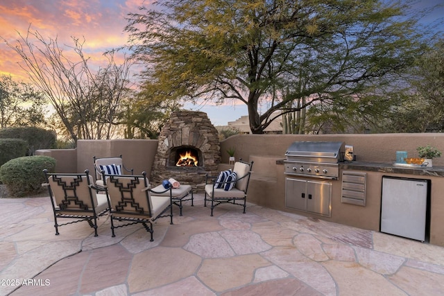 patio terrace at dusk featuring area for grilling, an outdoor stone fireplace, and an outdoor kitchen